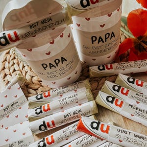 May include: A collection of small chocolate bars wrapped in red and white foil with various messages printed on them. The messages include "Du bist mein gr&ouml;&szlig;ter Fan" (You are my biggest fan), "Du bist einfach wunderbar" (You are simply wonderful), "Du bist ein tolles Vorbild" (You are a great role model), and "Du machst jeden Quatsch mit" (You do every silly thing with me).
