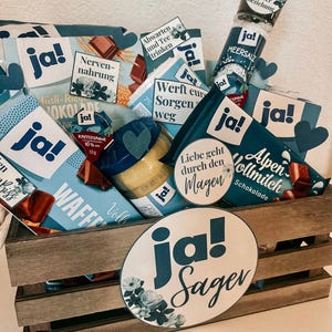 Puede incluir: Una caja de madera llena de diversos regalos y golosinas, incluyendo barras de chocolate, un tarro de crema de caf&eacute;, un tarro de mostaza, un tarro de sal marina y una peque&ntilde;a bolsa de t&eacute;. La caja est&aacute; decorada con c&iacute;rculos de papel azul y blanco que dicen "ja!" y "ja! Sager".