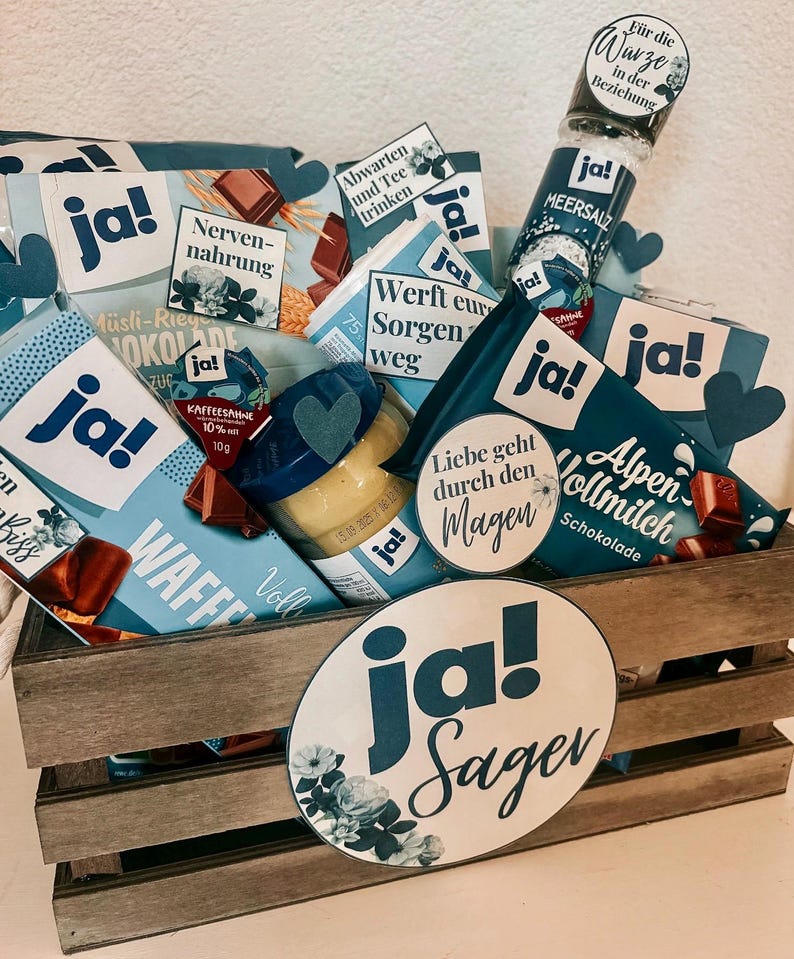Puede incluir: Una caja de madera llena de diversos regalos y golosinas, incluyendo barras de chocolate, un tarro de crema de caf&eacute;, un tarro de mostaza, un tarro de sal marina y una peque&ntilde;a bolsa de t&eacute;. La caja est&aacute; decorada con c&iacute;rculos de papel azul y blanco que dicen "ja!" y "ja! Sager".