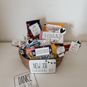Puede incluir: Una cesta tejida llena de aperitivos y peque&ntilde;os regalos, etiquetada como un "Kit de supervivencia para un nuevo trabajo". El kit incluye varias golosinas empaquetadas y tarjetas con frases alemanas.