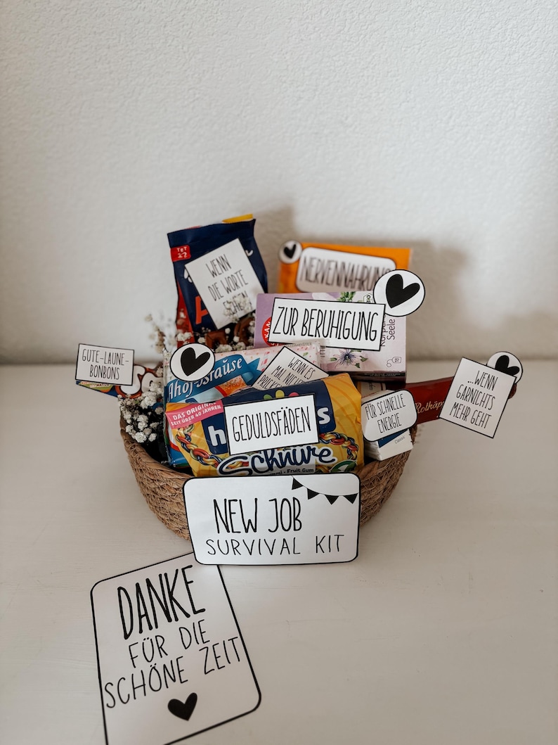 Puede incluir: Una cesta tejida llena de aperitivos y peque&ntilde;os regalos, etiquetada como un "Kit de supervivencia para un nuevo trabajo". El kit incluye varias golosinas empaquetadas y tarjetas con frases alemanas.