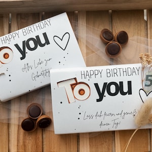 May include: Two white gift boxes with "Happy Birthday To You" text, a heart, and decorative dots. One box is tied with twine. Chocolate candies and dried flowers are also present. The boxes have German text.