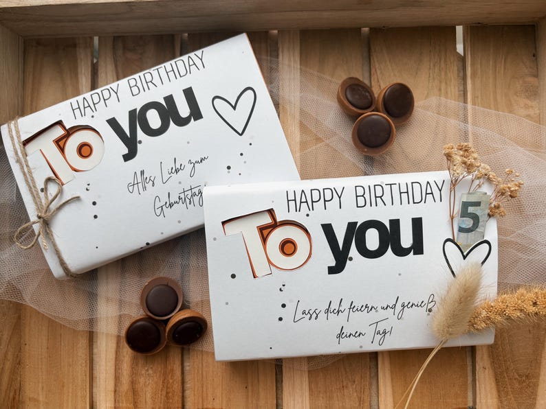 May include: Two white gift boxes with "Happy Birthday To You" text, a heart, and decorative dots. One box is tied with twine. Chocolate candies and dried flowers are also present. The boxes have German text.