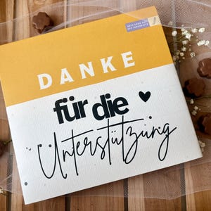 May include: A rectangular card with a yellow top section and a white bottom section. The word "DANKE" is printed in white block letters on the yellow section. The words "f&uuml;r die Unterst&uuml;tzung" are printed in black script on the white section.