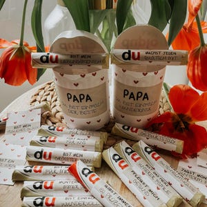 May include: A collection of small wrapped chocolate bars with the word "du" printed on each bar. The bars have different messages printed on them, such as "Bist mein gr&ouml;&szlig;ter Fan" and "Gibst immer 150 Prozent". There are two white cups with the word "Papa" printed on them. The cups are decorated with red hearts. The cups are sitting on a wooden surface with a bouquet of red tulips in the background.