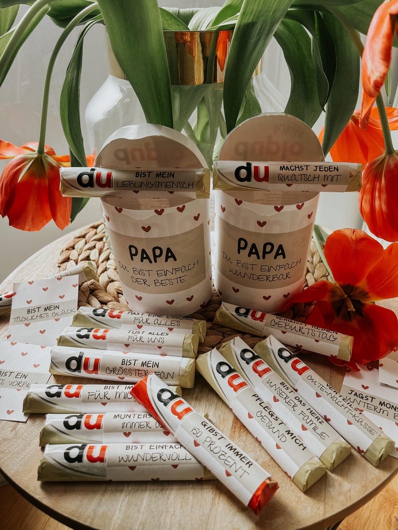 May include: A collection of small wrapped chocolate bars with the word "du" printed on each bar. The bars have different messages printed on them, such as "Bist mein gr&ouml;&szlig;ter Fan" and "Gibst immer 150 Prozent". There are two white cups with the word "Papa" printed on them. The cups are decorated with red hearts. The cups are sitting on a wooden surface with a bouquet of red tulips in the background.