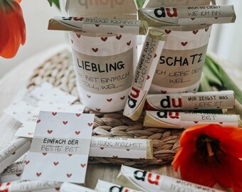 May include: Two white cups with red heart patterns and German text, filled with rolled-up messages. The cups are labeled "Liebling" and "Schatz." Scattered around are more messages and a few red tulips.