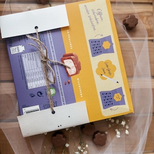 May include: A box of Milka pralines with a purple and yellow design, tied with twine. The box features text and illustrations, with a focus on the product's milk cream filling. The background includes chocolate flower-shaped candies and delicate white flowers.