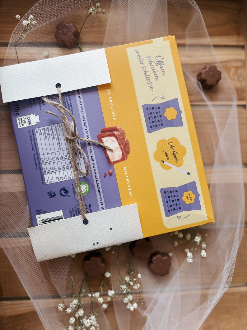 May include: A box of Milka pralines with a purple and yellow design, tied with twine. The box features text and illustrations, with a focus on the product's milk cream filling. The background includes chocolate flower-shaped candies and delicate white flowers.
