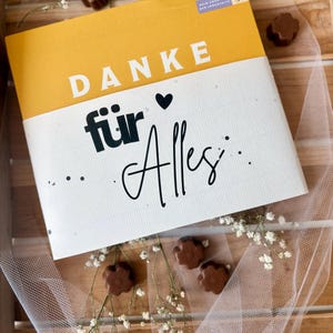 May include: A greeting card with the words "DANKE f&uuml;r Alles" in black and white, with a yellow top section. The card is decorated with small chocolate candies and white flowers, arranged on a wooden surface.