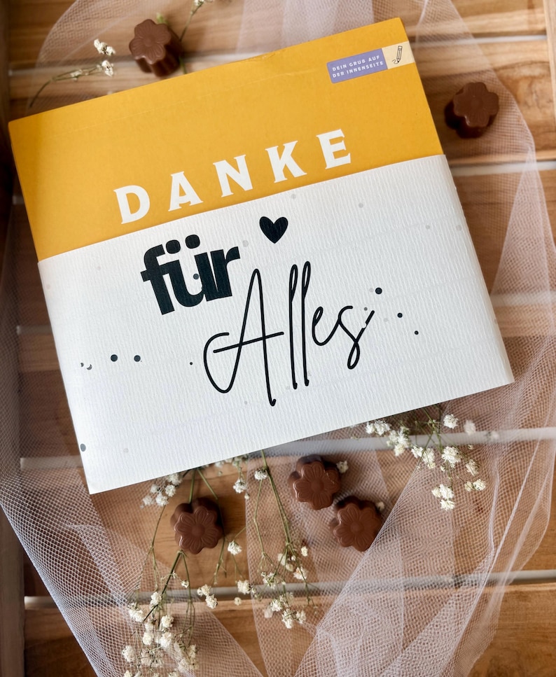 May include: A greeting card with the words "DANKE f&uuml;r Alles" in black and white, with a yellow top section. The card is decorated with small chocolate candies and white flowers, arranged on a wooden surface.