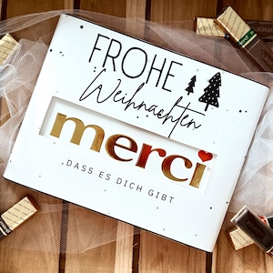 Op de afbeelding: Een witte doos met de woorden "FROHE Weihnachten" en "merci" in gouden letters. De doos heeft een venster dat het woord "merci" en een klein rood hart onthult. Chocoladerepen liggen verspreid rond de doos.