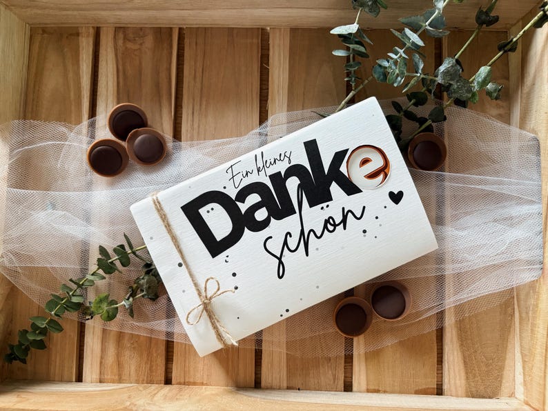 May include: A white rectangular gift box with the words "Danke Sch&ouml;n" in black script and a small heart. The box is tied with twine and surrounded by chocolate candies and greenery, all arranged on a wooden tray.