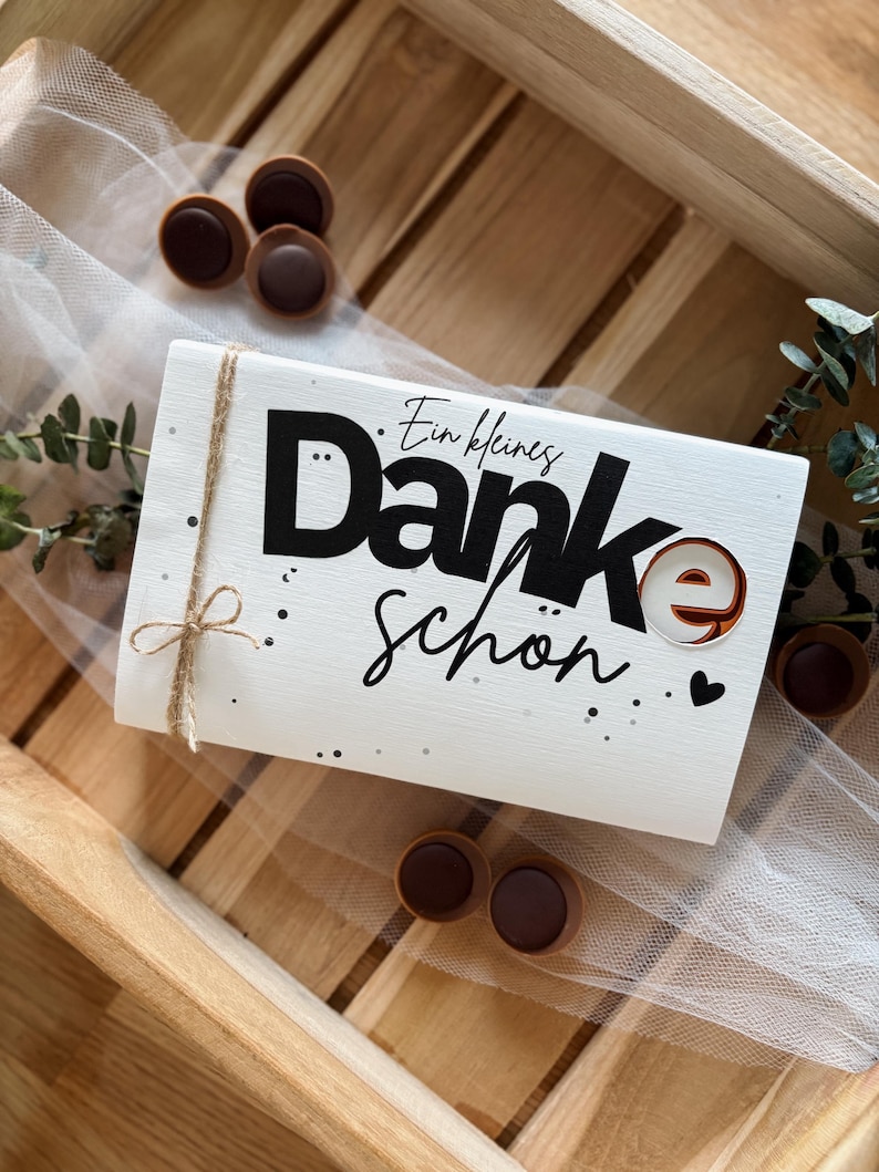 May include: A white rectangular gift box with the words "Danke sch&ouml;n" in black script and a brown candy accent. The box is tied with twine and surrounded by chocolate candies and greenery.