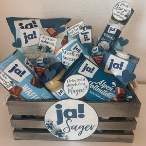 Puede incluir: Una caja de madera llena de varios art&iacute;culos de comida y bebida, incluyendo chocolate, crema de caf&eacute; y un tarro de miel. La caja est&aacute; decorada con c&iacute;rculos de papel azul y blanco que dicen "ja!" y "Sager".