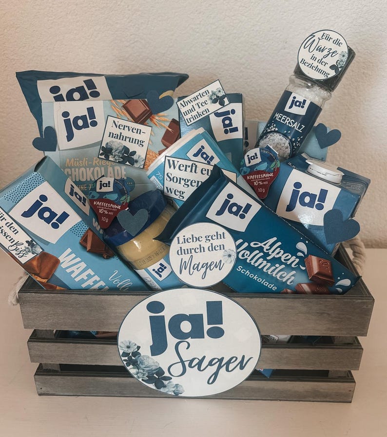 Puede incluir: Una caja de madera llena de varios art&iacute;culos de comida y bebida, incluyendo chocolate, crema de caf&eacute; y un tarro de miel. La caja est&aacute; decorada con c&iacute;rculos de papel azul y blanco que dicen "ja!" y "Sager".