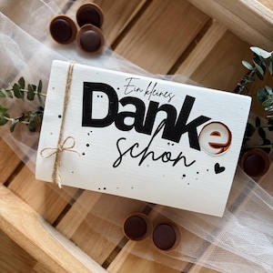 May include: A white rectangular gift box with the German words "Ein kleines Danke sch&ouml;n" in black script and a brown accent. The box is tied with twine and surrounded by chocolate candies.