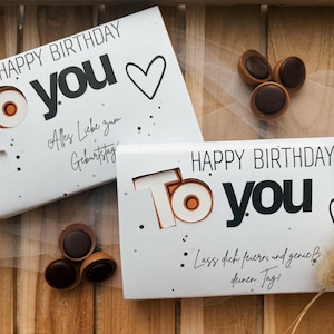 May include: Two white gift boxes with "HAPPY BIRTHDAY TO YOU" text, a heart, and decorative accents. The boxes are tied with twine and accompanied by chocolate candies. The image is set on a wooden surface.