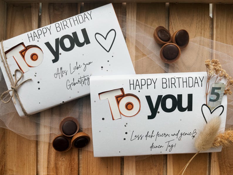 May include: Two white gift boxes with "HAPPY BIRTHDAY TO YOU" text, a heart, and decorative accents. The boxes are tied with twine and accompanied by chocolate candies. The image is set on a wooden surface.