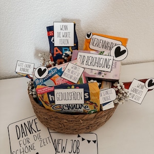 Puede incluir: Una cesta de mimbre llena de aperitivos y golosinas, incluyendo caramelos y patatas fritas. La cesta tiene varios letreros peque&ntilde;os con texto en alem&aacute;n, incluyendo "New Job Survival Kit" y "Danke f&uuml;r die sch&ouml;ne Zeit."