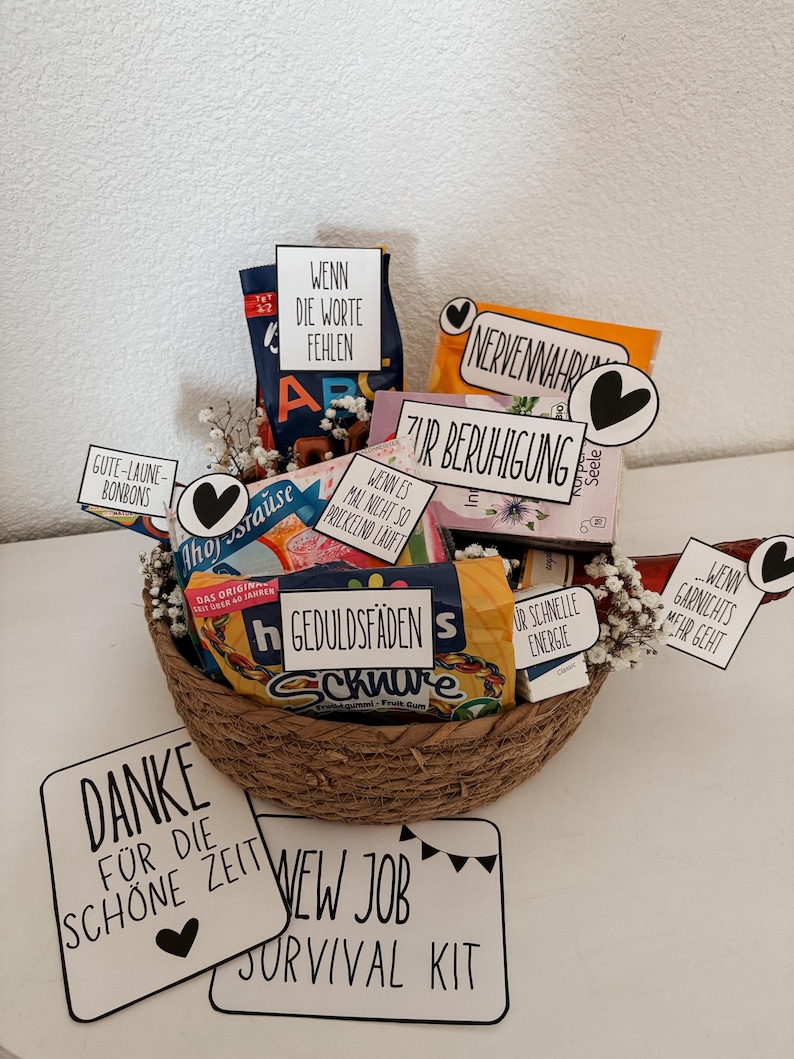 Puede incluir: Una cesta de mimbre llena de aperitivos y golosinas, incluyendo caramelos y patatas fritas. La cesta tiene varios letreros peque&ntilde;os con texto en alem&aacute;n, incluyendo "New Job Survival Kit" y "Danke f&uuml;r die sch&ouml;ne Zeit."