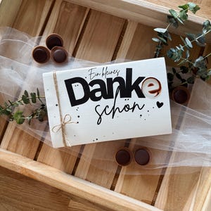 May include: A white gift box with the words "Danke sch&ouml;n" in black script and a small heart. The box is tied with twine and sits on a wooden tray with chocolate candies and greenery.