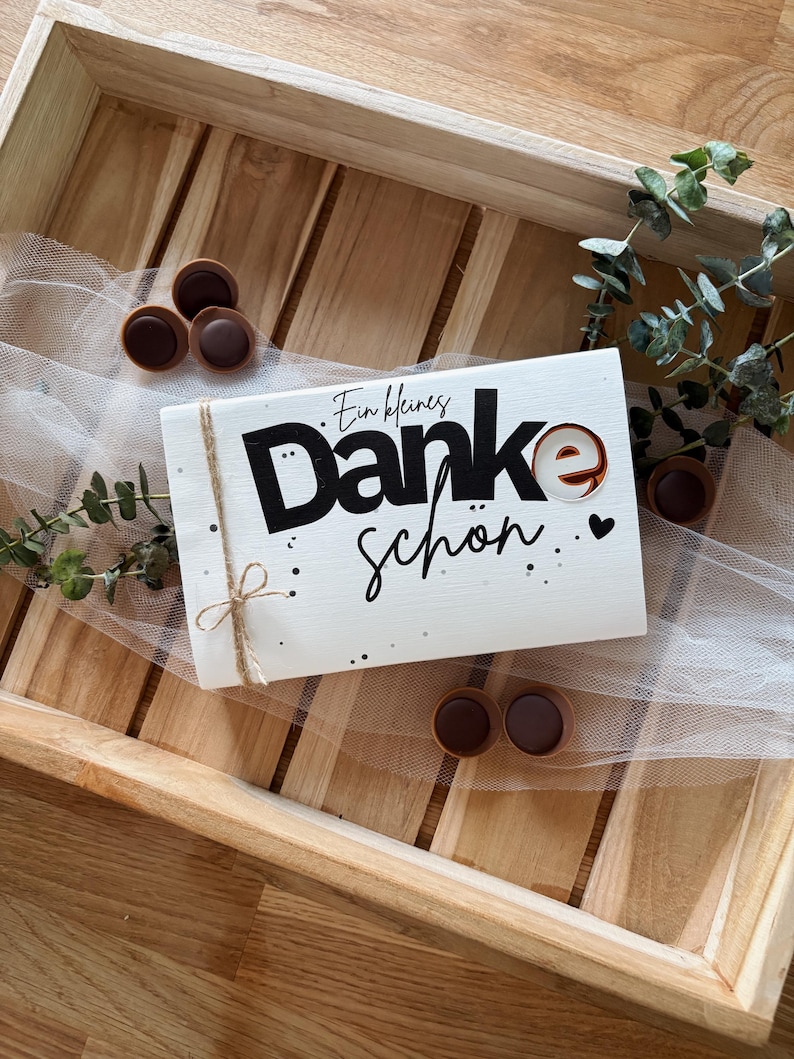 May include: A white gift box with the words "Danke sch&ouml;n" in black script and a small heart. The box is tied with twine and sits on a wooden tray with chocolate candies and greenery.