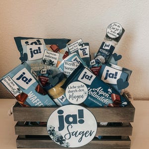 Puede incluir: Una caja de madera llena de varios art&iacute;culos de comida, incluyendo chocolate, dulces y un tarro de sal. La caja est&aacute; decorada con c&iacute;rculos de papel azul y blanco que dicen "ja!" y "ja! Sager".