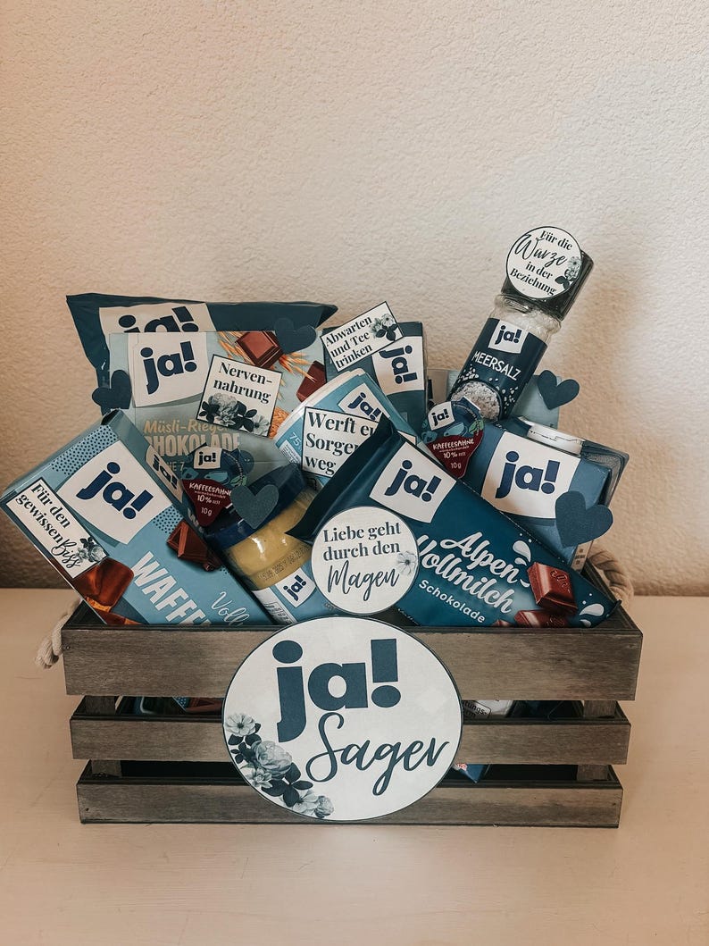 Puede incluir: Una caja de madera llena de varios art&iacute;culos de comida, incluyendo chocolate, dulces y un tarro de sal. La caja est&aacute; decorada con c&iacute;rculos de papel azul y blanco que dicen "ja!" y "ja! Sager".