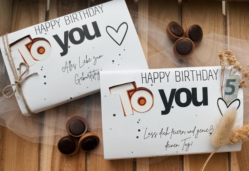 May include: Two white gift boxes with "HAPPY BIRTHDAY TO YOU" text, a heart, and decorative dots. One box is tied with twine. Chocolate candies are scattered around the boxes. The boxes are on a wooden surface.