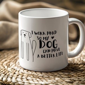Könnte beinhalten: Weiße Keramik-Tasse mit einer schwarzen Strichzeichnung eines Hundes und dem Text "I WORK HARD SO MY DOG CAN HAVE A BETTER LIFE." Die Tasse hat einen gebogenen Henkel und steht auf einer gewebten Matte.