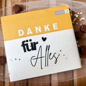 May include: A greeting card with the words "DANKE f&uuml;r Alles" in black script and a yellow and white color scheme. The card is decorated with small chocolate candies and sprigs of white flowers.