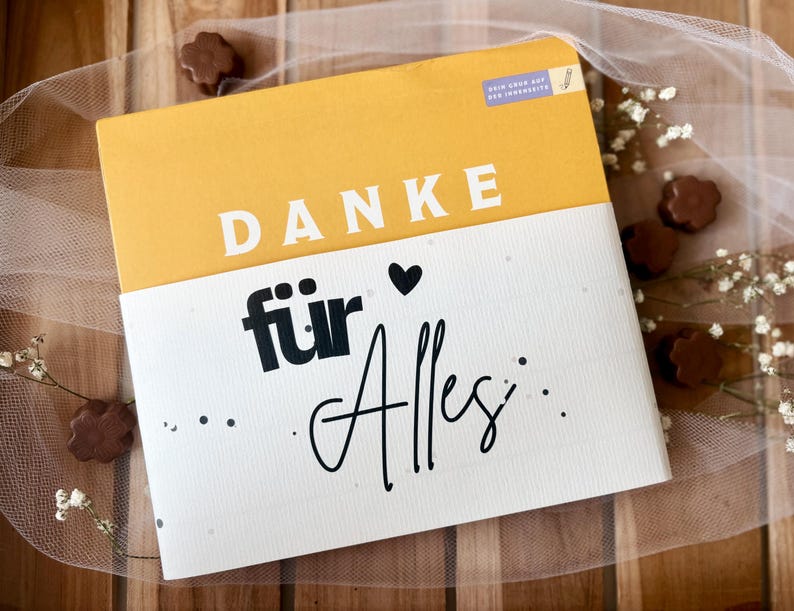 May include: A greeting card with the words "DANKE f&uuml;r Alles" in black script and a yellow and white color scheme. The card is decorated with small chocolate candies and sprigs of white flowers.
