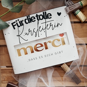 May include: A white gift box with gold lettering that says "merci" in a window. The box has the text "Für die tolle Kursleiterin, dass es dich gibt" in black script. Several chocolate bars are scattered around the box.