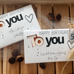 May include: Two white gift boxes with "HAPPY BIRTHDAY TO YOU" printed in black and copper. One box is tied with twine. The boxes are decorated with hearts and dots. Chocolate candies are scattered around the boxes.