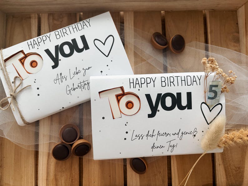 May include: Two white gift boxes with "HAPPY BIRTHDAY TO YOU" printed in black and copper. One box is tied with twine. The boxes are decorated with hearts and dots. Chocolate candies are scattered around the boxes.