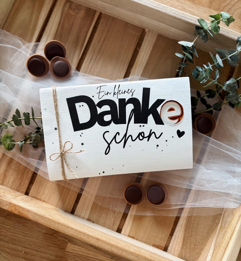 May include: A white wooden sign with the words "Ein kleines Danke sch&ouml;n" in black script and a decorative "e". The sign is tied with twine and decorated with small black dots. Several chocolate candies are scattered around the sign.
