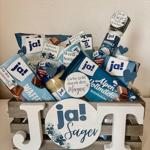 Puede incluir: Una caja de madera llena de varios regalos de tem&aacute;tica azul y blanca, incluyendo barras de chocolate, un tarro de miel y una peque&ntilde;a botella de sal marina. La caja est&aacute; decorada con una gran letra J de madera blanca y una pegatina redonda azul y blanca que dice "Ja! Sager".