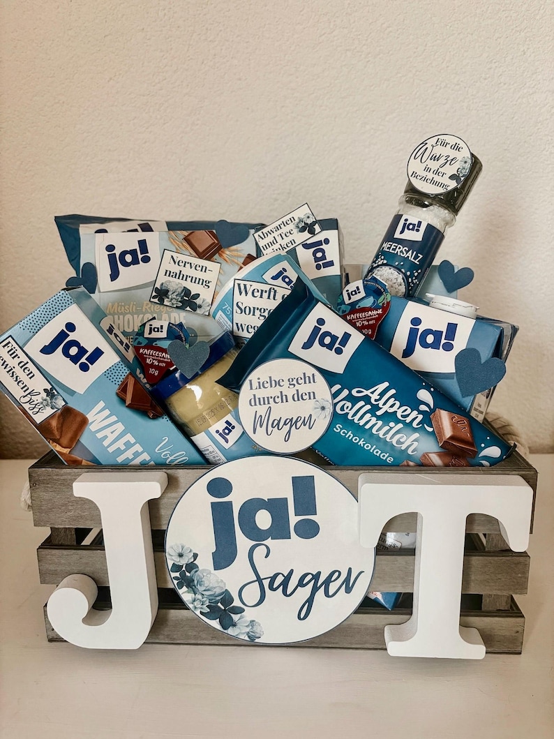 Puede incluir: Una caja de madera llena de varios regalos de tem&aacute;tica azul y blanca, incluyendo barras de chocolate, un tarro de miel y una peque&ntilde;a botella de sal marina. La caja est&aacute; decorada con una gran letra J de madera blanca y una pegatina redonda azul y blanca que dice "Ja! Sager".