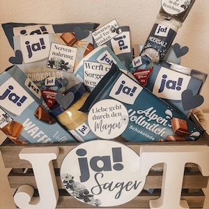 Puede incluir: Una caja de madera llena de varios art&iacute;culos de comida, incluyendo barras de chocolate, un tarro de sal marina y una botella de jarabe. La caja est&aacute; decorada con c&iacute;rculos de papel azul y blanco que dicen "ja!" y "ja! sagen".
