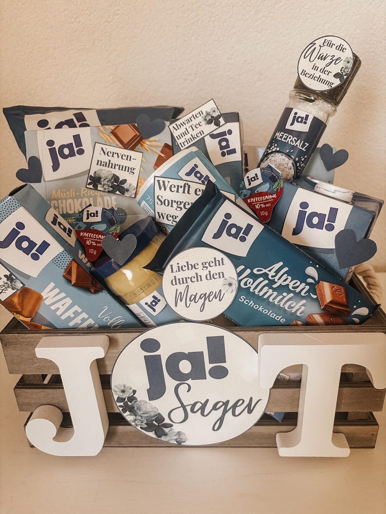 Puede incluir: Una caja de madera llena de varios art&iacute;culos de comida, incluyendo barras de chocolate, un tarro de sal marina y una botella de jarabe. La caja est&aacute; decorada con c&iacute;rculos de papel azul y blanco que dicen "ja!" y "ja! sagen".