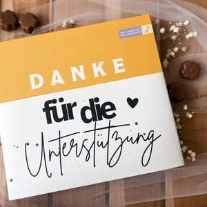 May include: A greeting card with the German word "DANKE" in white block letters on a yellow background. Below, the words "f&uuml;r die Unterst&uuml;tzung" are written in black script. A black heart is next to the word "die".