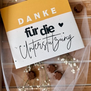 May include: A notebook with a yellow and white cover, featuring the German text "DANKE f&uuml;r die Unterst&uuml;tzung" in bold and script fonts. The image includes small chocolate flower-shaped candies and sprigs of white flowers.