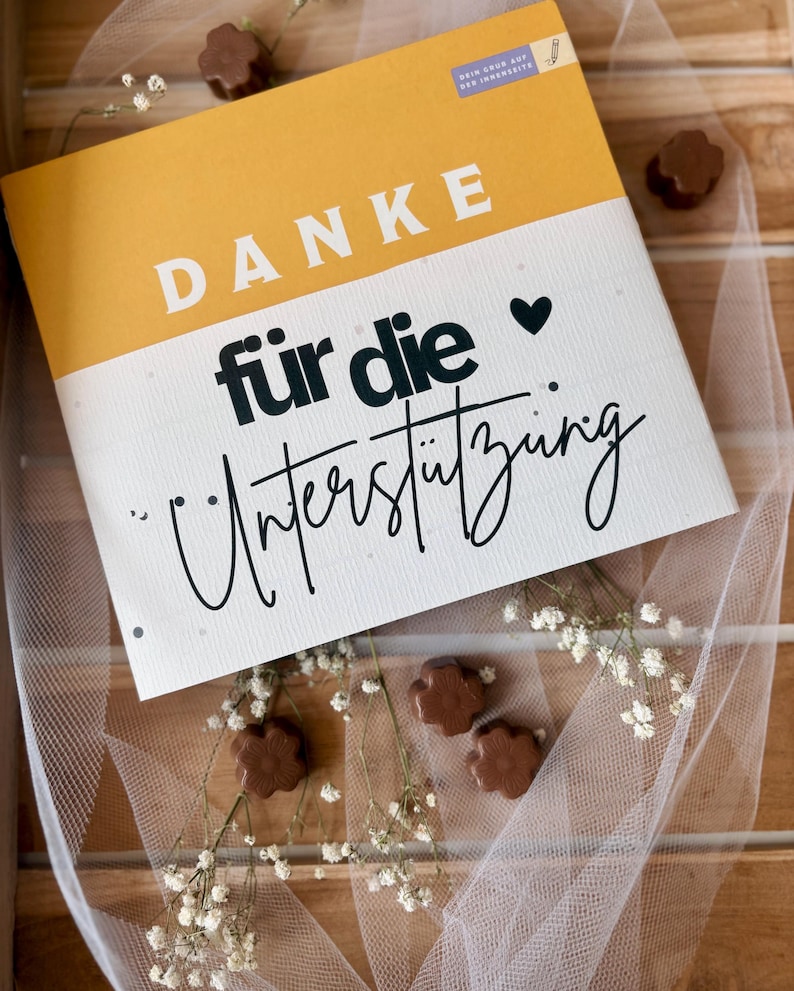 May include: A notebook with a yellow and white cover, featuring the German text "DANKE f&uuml;r die Unterst&uuml;tzung" in bold and script fonts. The image includes small chocolate flower-shaped candies and sprigs of white flowers.