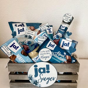 Puede incluir: Una caja de madera llena de varios art&iacute;culos de regalo azules y blancos, incluyendo barras de chocolate, un tarro de mantequilla de man&iacute;, un tarro de sal marina y una peque&ntilde;a bolsa de regalo azul y blanca. La caja est&aacute; decorada con un c&iacute;rculo grande azul y blanco con el texto "ja! Sager" en blanco.