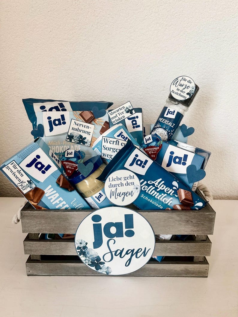 Puede incluir: Una caja de madera llena de varios art&iacute;culos de regalo azules y blancos, incluyendo barras de chocolate, un tarro de mantequilla de man&iacute;, un tarro de sal marina y una peque&ntilde;a bolsa de regalo azul y blanca. La caja est&aacute; decorada con un c&iacute;rculo grande azul y blanco con el texto "ja! Sager" en blanco.