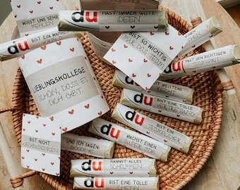 Puede incluir: Un contenedor cilíndrico blanco con un patrón de corazones rojos y el texto "Liebling-Kollege Schön, dass es dich gibt." El contenedor está lleno de pequeñas barras de chocolate envueltas en papel blanco con la palabra "du" impresa en rojo. Cada barra de chocolate tiene un mensaje diferente impreso en ella, como "Bist so wichtig für das Team" y "Hast den richtigen Teamspirit".