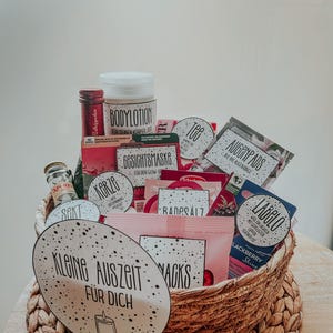 May include: A wicker basket filled with various self-care items, including bath salts, body lotion, face masks, tea, and a candle. The basket is decorated with a tag that reads "Kleine Auszeit f&uuml;r Dich" which translates to "Small Timeout for You".