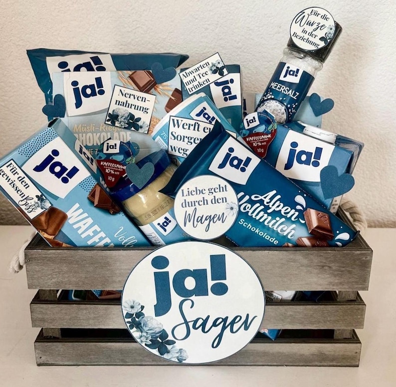 Puede incluir: Una cesta de regalo azul y blanca llena de varios art&iacute;culos de comida, incluyendo chocolate, obleas y un tarro de mantequilla de cacahuete. La cesta est&aacute; decorada con papel azul y blanco y un c&iacute;rculo grande azul y blanco con el texto "ja! Sager".