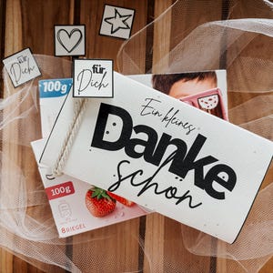 以下が含まれることがあります： 黒いスクリプトで「Ein kleines Danke schön」というドイツ語のテキストが入った白いギフトバッグと、小さな装飾タグ。画像にはイチゴの箱と、透き通った白い生地が含まれています。